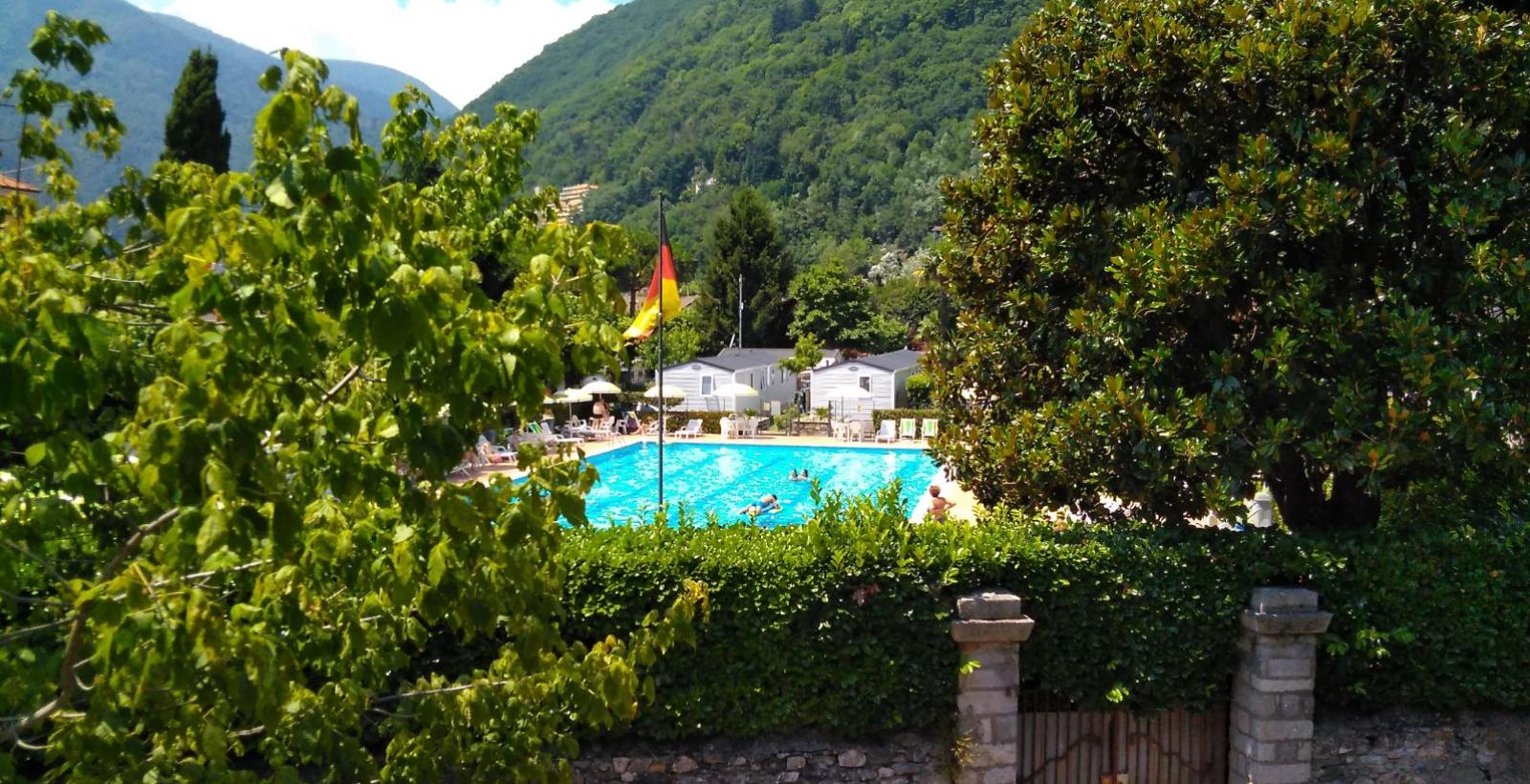 Piscina all'aperto circondata da montagne e verde, con bandiera visibile.