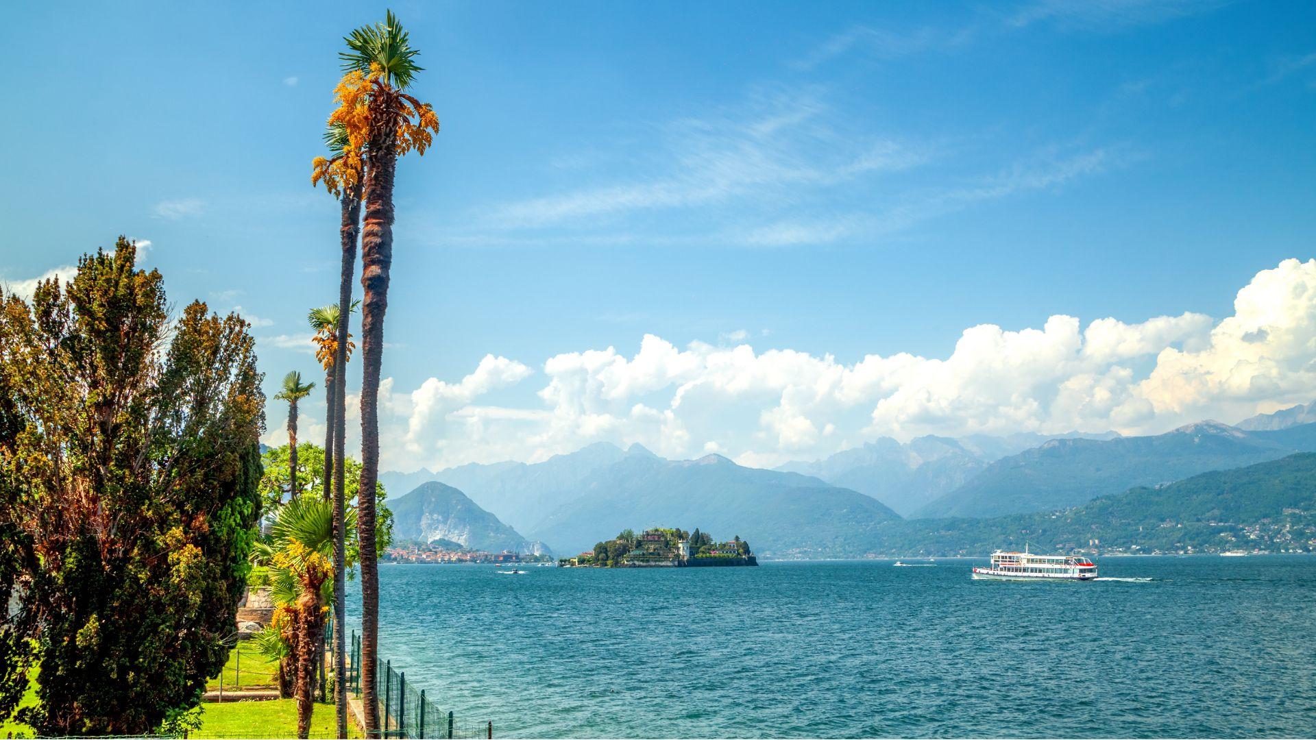 Panorama lacustre con palme, isola e barca, circondato da montagne e cielo azzurro.