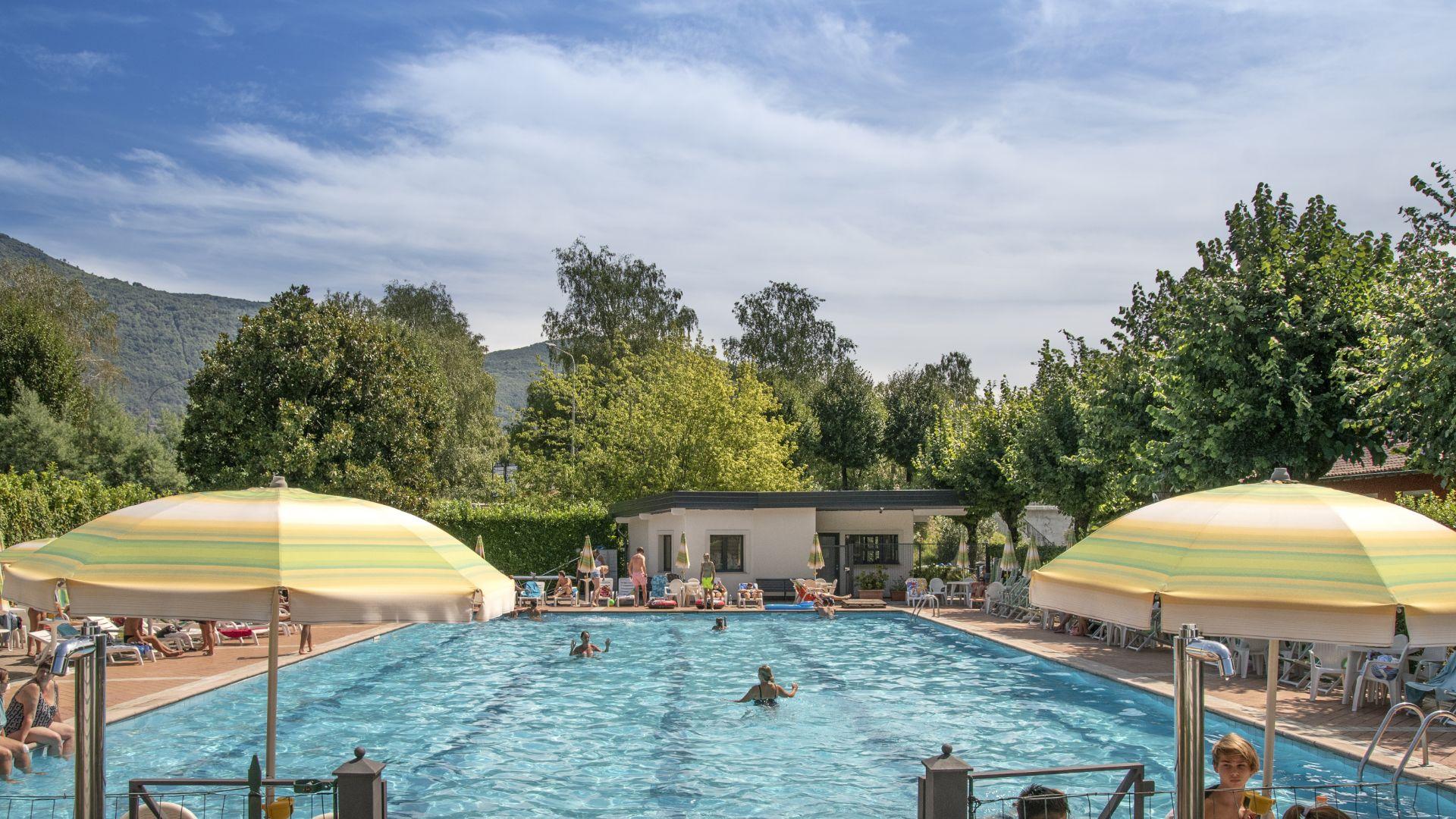 Piscina all'aperto con ombrelloni gialli, circondata da alberi e montagne.