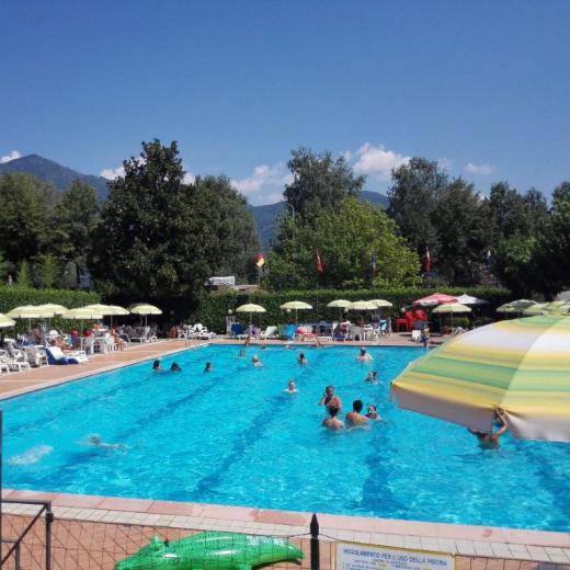 Piscina all'aperto con bagnanti, ombrelloni e montagne sullo sfondo.