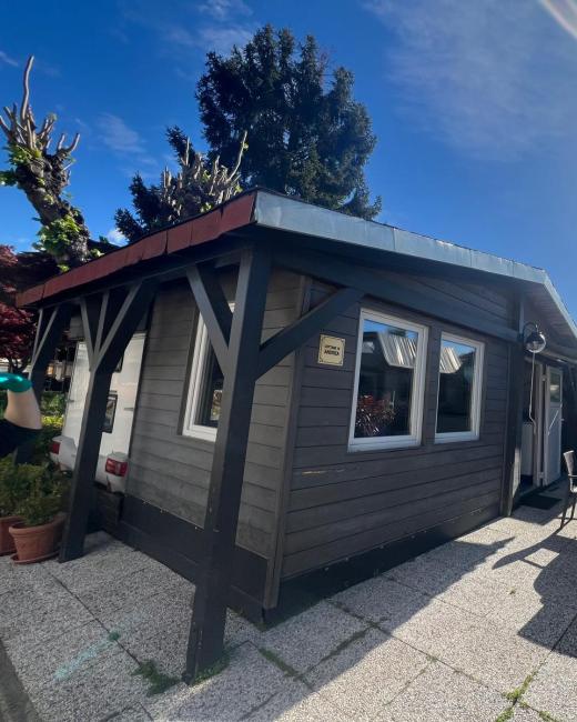 Casetta in legno con terrazza soleggiata, circondata da piante e cielo sereno.