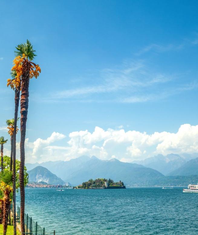 Paesaggio lacustre con palme, isola e barca, circondato da montagne e cielo azzurro.