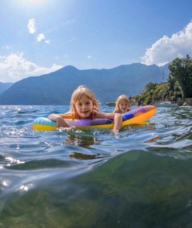 Bambine giocano in acqua con un materassino, montagne e cielo sereno sullo sfondo.