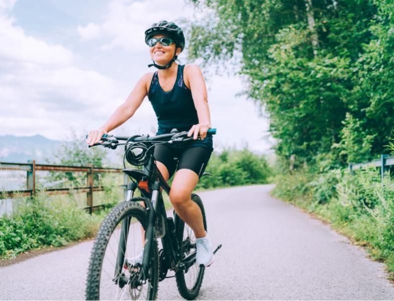 Donna in bicicletta su pista ciclabile immersa nella natura, sorridente e attrezzata.