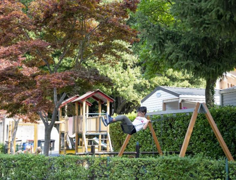 Bambino su altalena in parco giochi, circondato da alberi e casette.