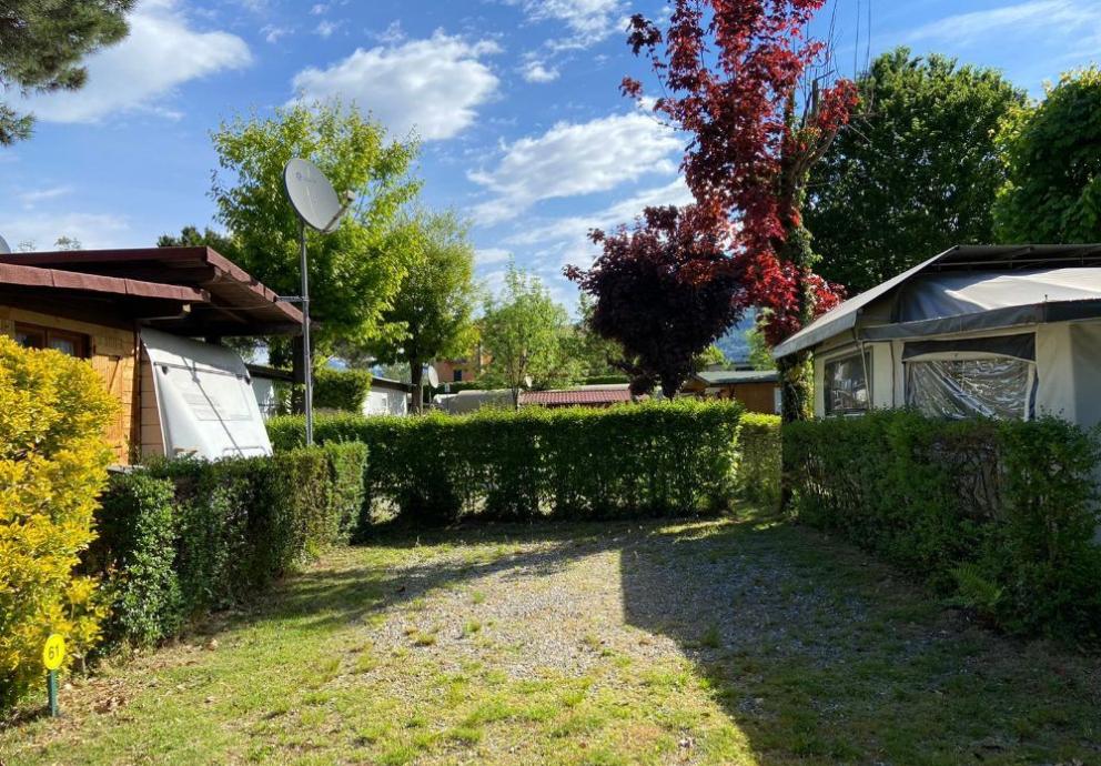 Area campeggio con roulotte, alberi e cielo sereno, ombreggiata da siepi verdi.