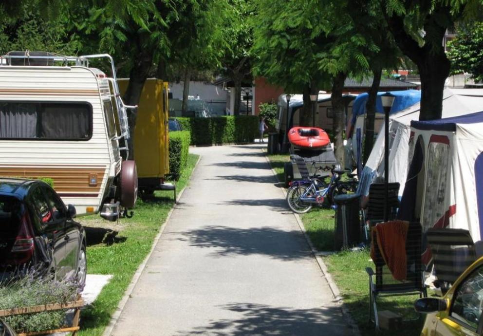 Campeggio con roulotte, tende, biciclette e un gommone lungo un viale alberato.